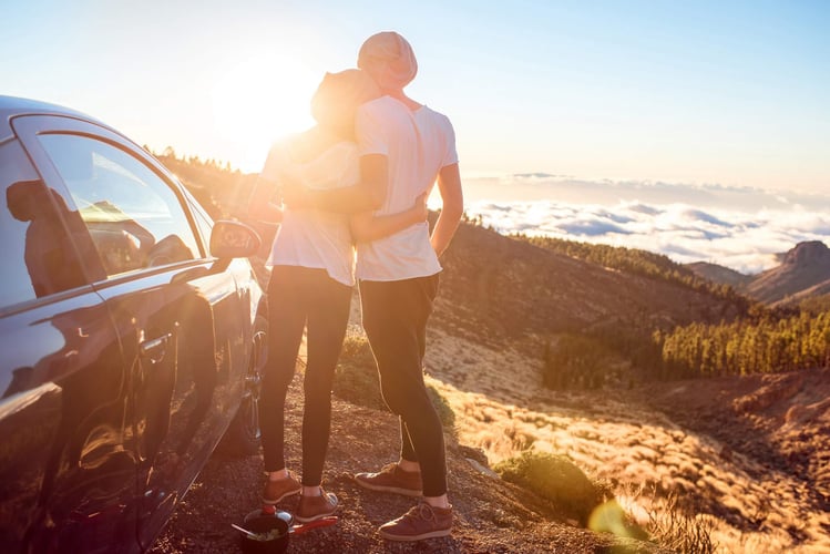 Image shows a couple embracing from behind beside a car with mountains and clouds ahead of them
