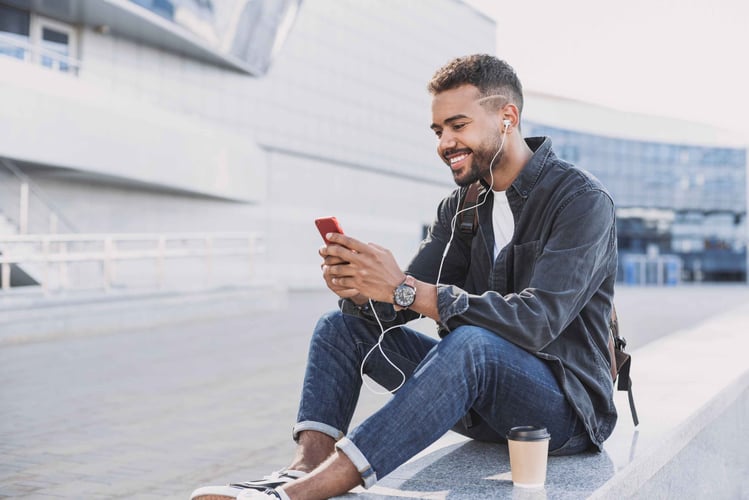 Image shows smiling young man sitting on a wall with earbuds in and looking at his phone screen