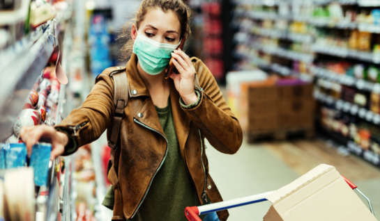 Image shows young woman in a mask behind a trolley in the aisle of a store lifting a blue box off a shelf