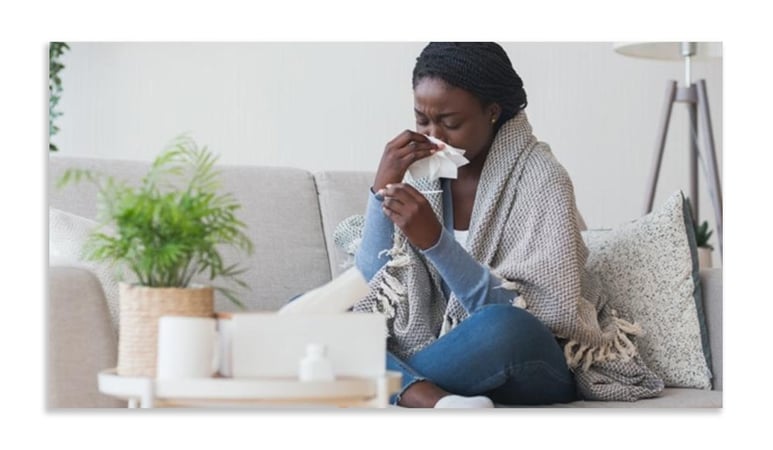 Image shows young woman sitting cross-legged on a sofa with a shawl around her shoulders and holding a tissue to her nose