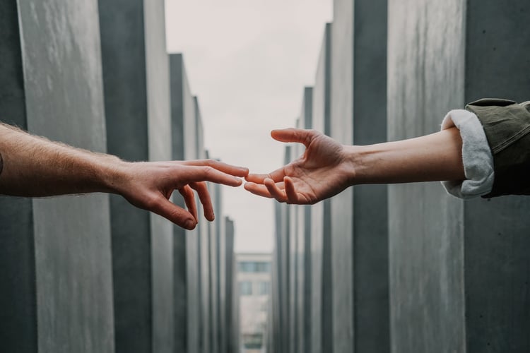 Two people's hands reaching towards each other with index fingers touching over the gulf between two buildings