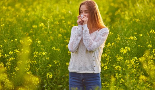 Image shows young woman in a field of yellow cowslips holding a tissue to her nose