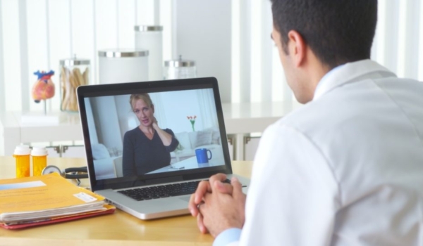 Image shows a man in a white coat from behind facing a laptop with a woman on screen holding her neck