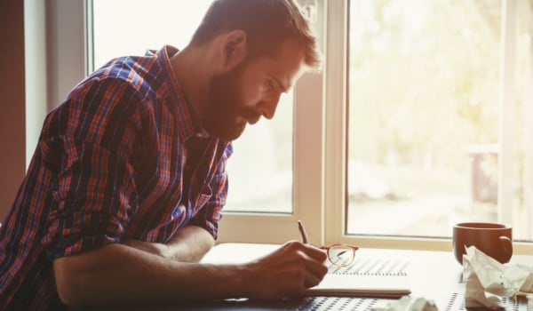 Images shows bearded man with pen in hand bent over a notepad at a table beside a window